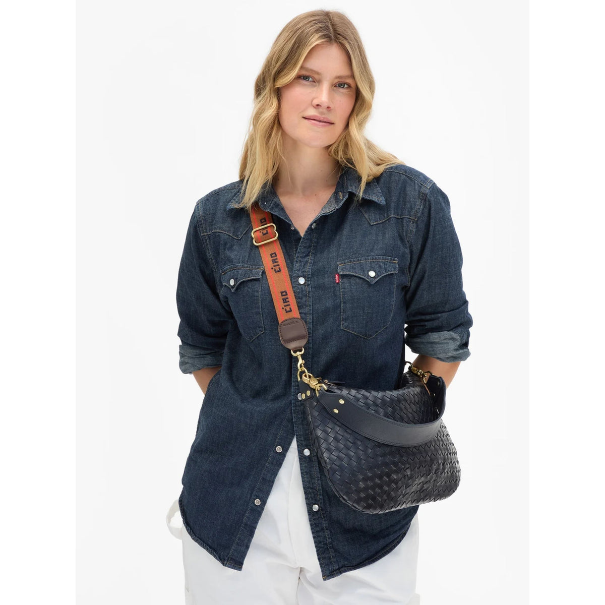Front view of a model wearing a dark denim button-up shirt and styling a woven black shoulder bag using the tan and orange "CIAO" printed adjustable crossbody strap.