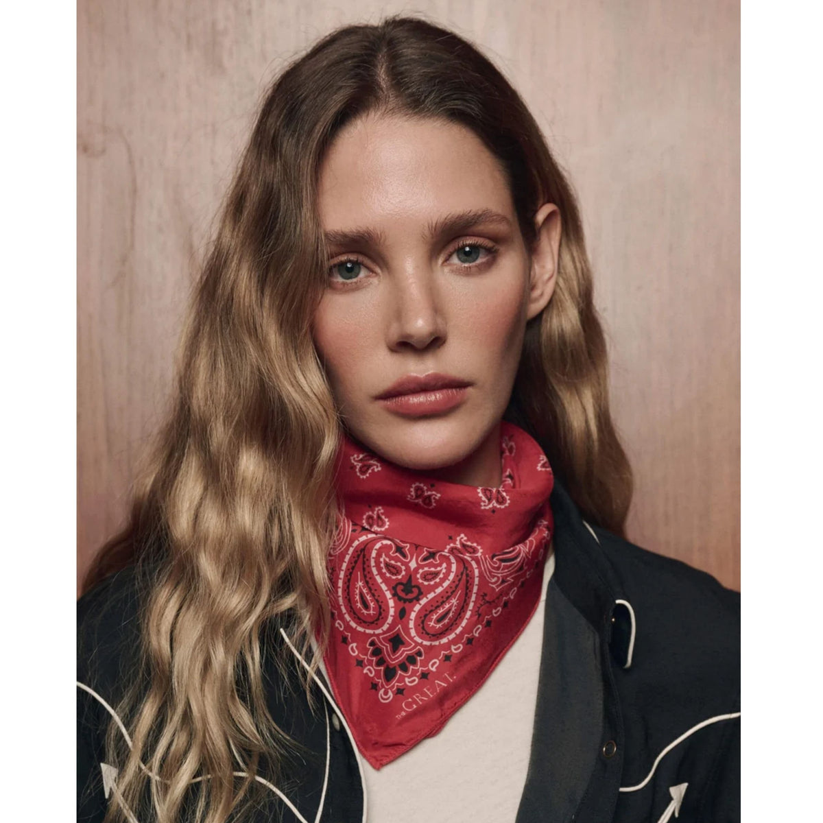 A woman with long wavy blonde hair wears a red paisley bandana around her neck, styled with a black jacket over a white shirt, set against a warm wood-paneled background.