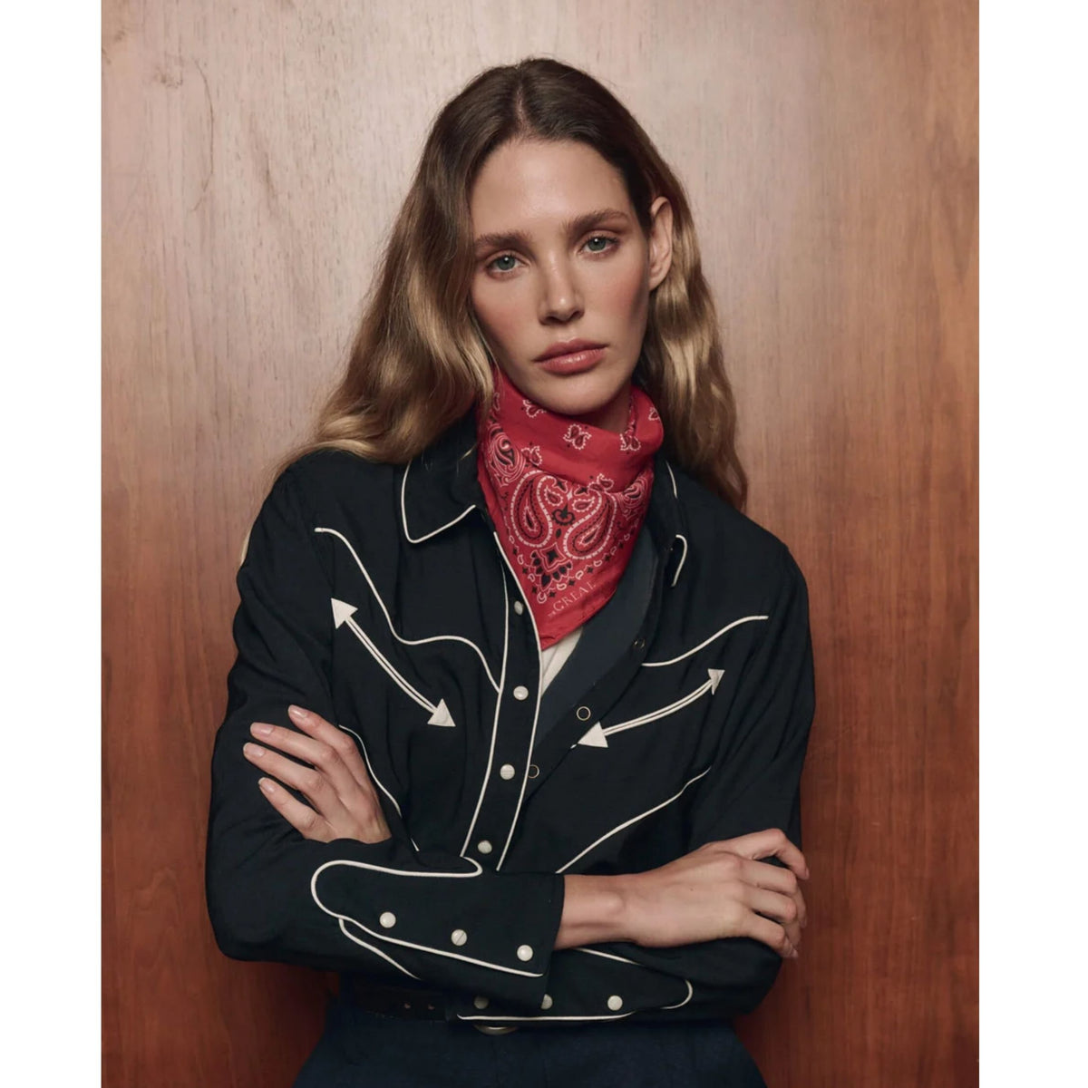 A woman with blonde-highlighted hair stands against a warm wood-paneled background, wearing a black western-style jacket with white piping and a red paisley bandana around her neck.