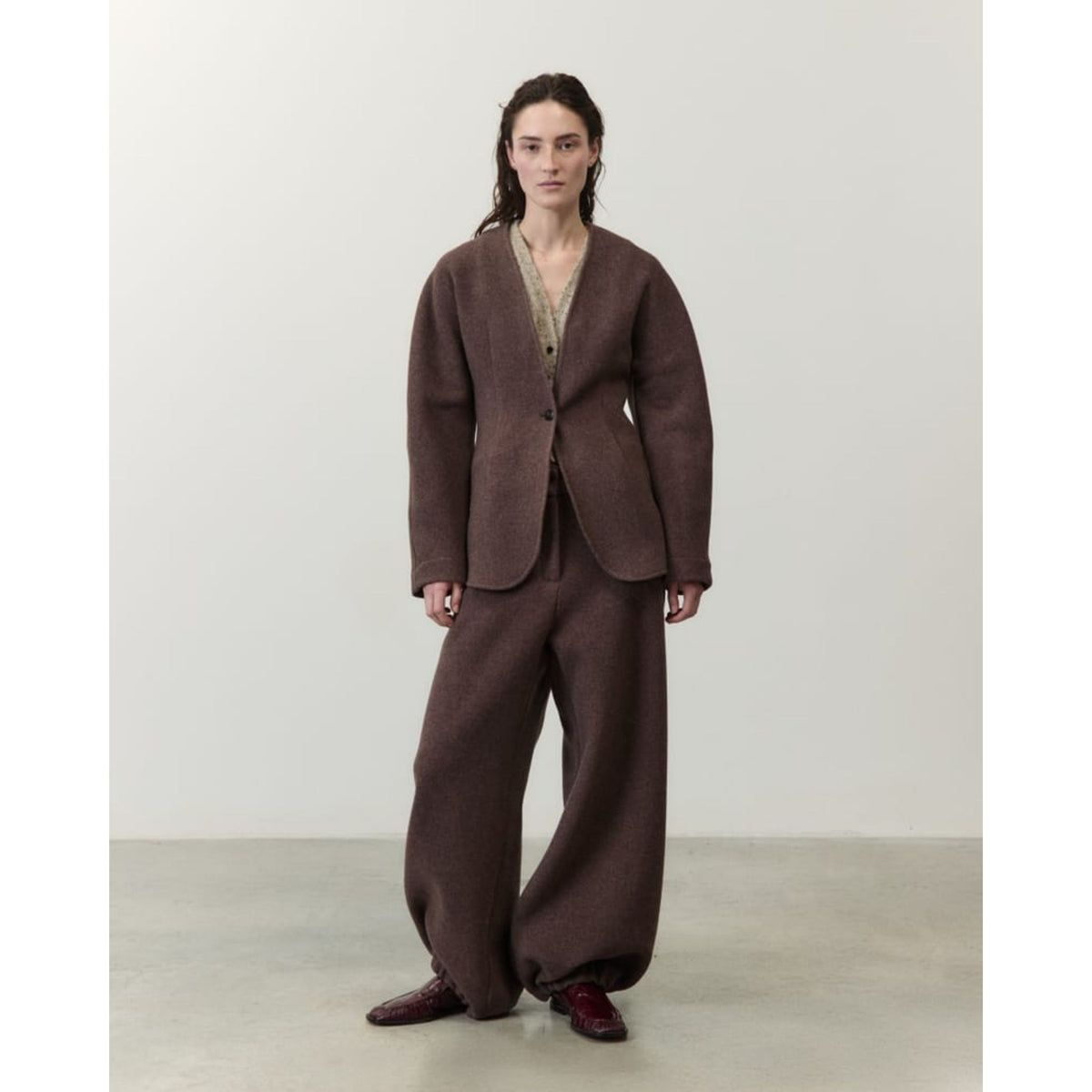 A model stands in a minimalist studio wearing a matching brown two-piece suit consisting of a textured open-front jacket and wide-leg trousers, paired with dark brown shoes.