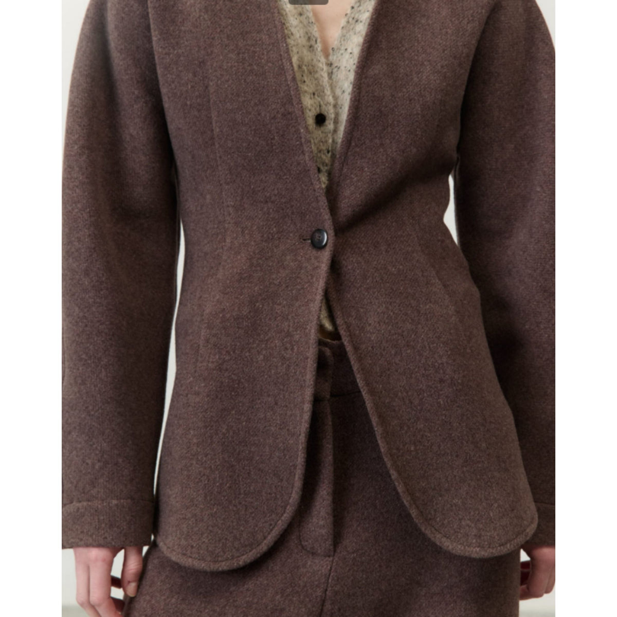 A close-up of a person wearing a brown wool-blend blazer with a single dark button, layered over a beige patterned knit top. The image is cropped from the neck down, showing the hands at the sides.