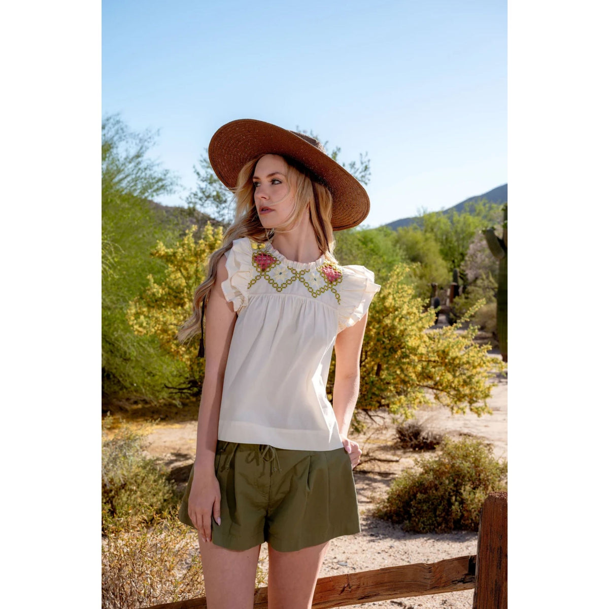 A woman stands outdoors in a desert landscape wearing a white sleeveless blouse with floral embroidery and ruffled shoulders, olive green shorts, and a wide brown hat.