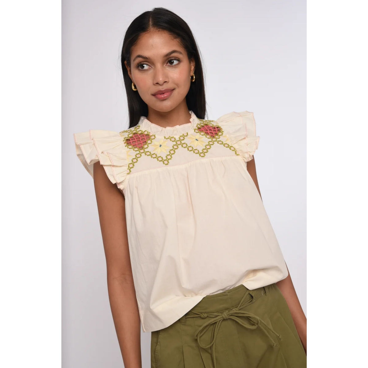 A woman wearing a cream-colored sleeveless blouse with ruffled shoulders and colorful floral embroidery across the chest, paired with olive green drawstring pants, set against a plain light gray background.