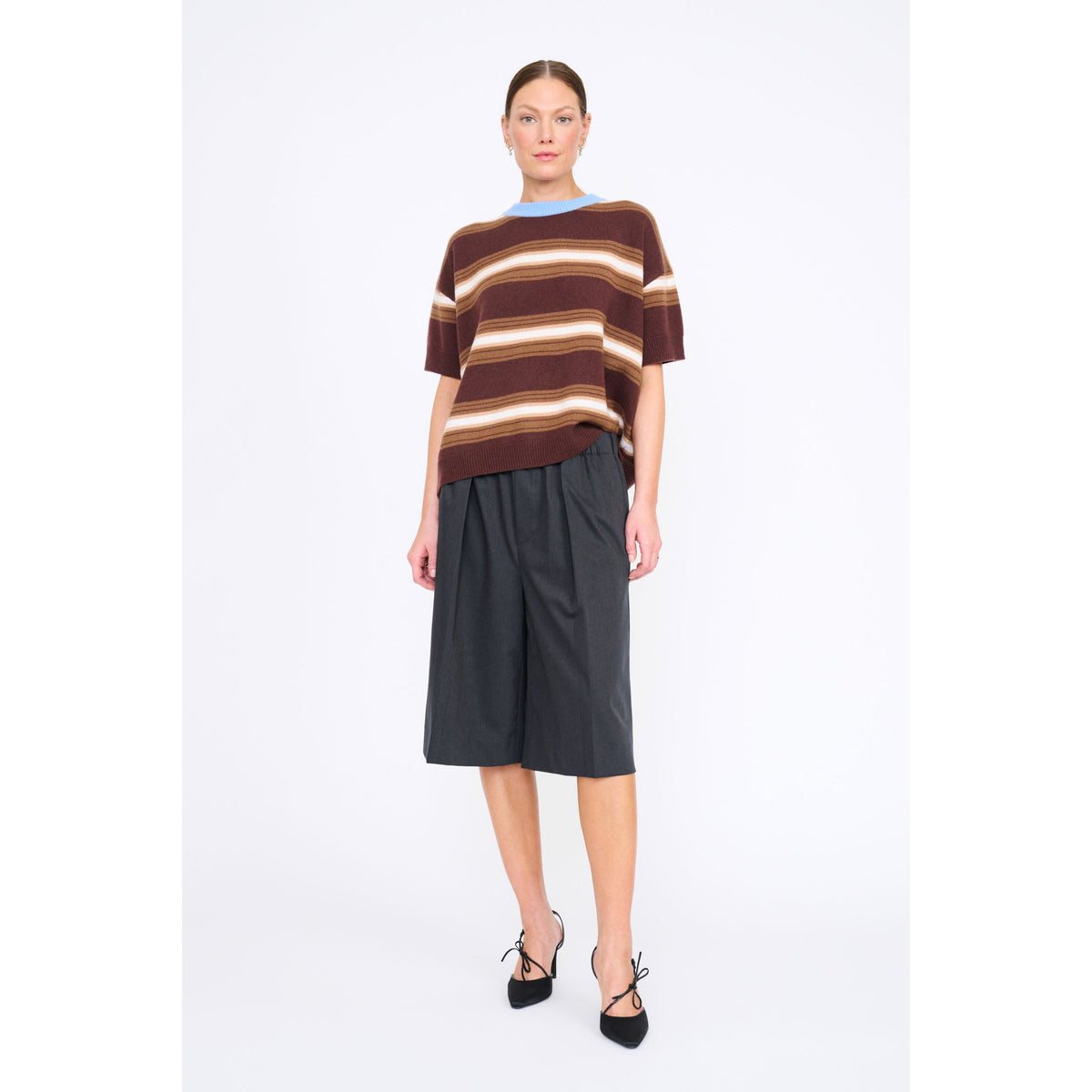 A model stands against a light gray studio background wearing a brown, beige, and white striped short-sleeve knit top, dark charcoal gray pleated shorts, and black lace-up heels.