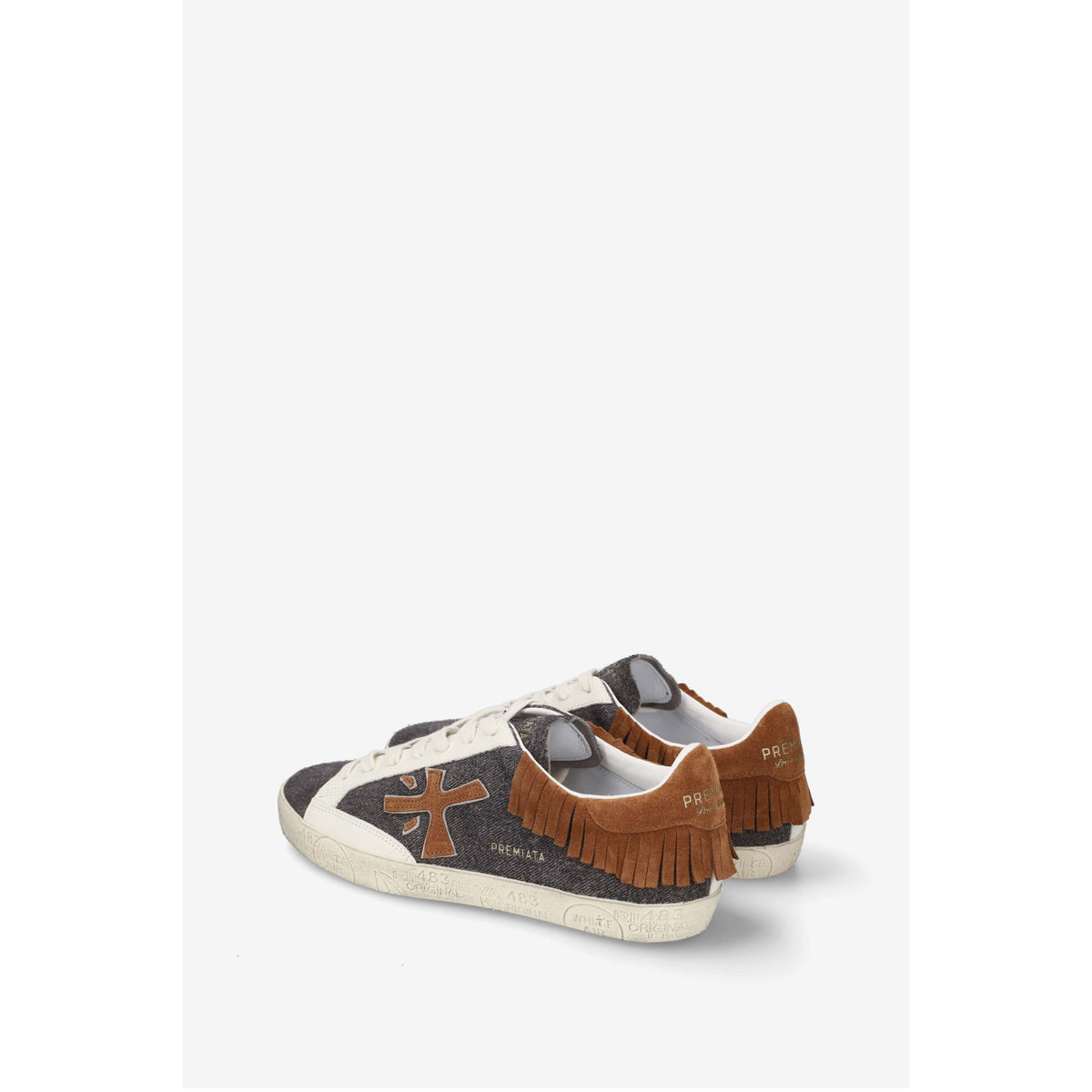 A pair of vintage-style low-top sneakers by PREMIATA, featuring a dark gray woven canvas upper, brown suede fringe on the heel, a brown cross logo on the side, and a white rubber sole.