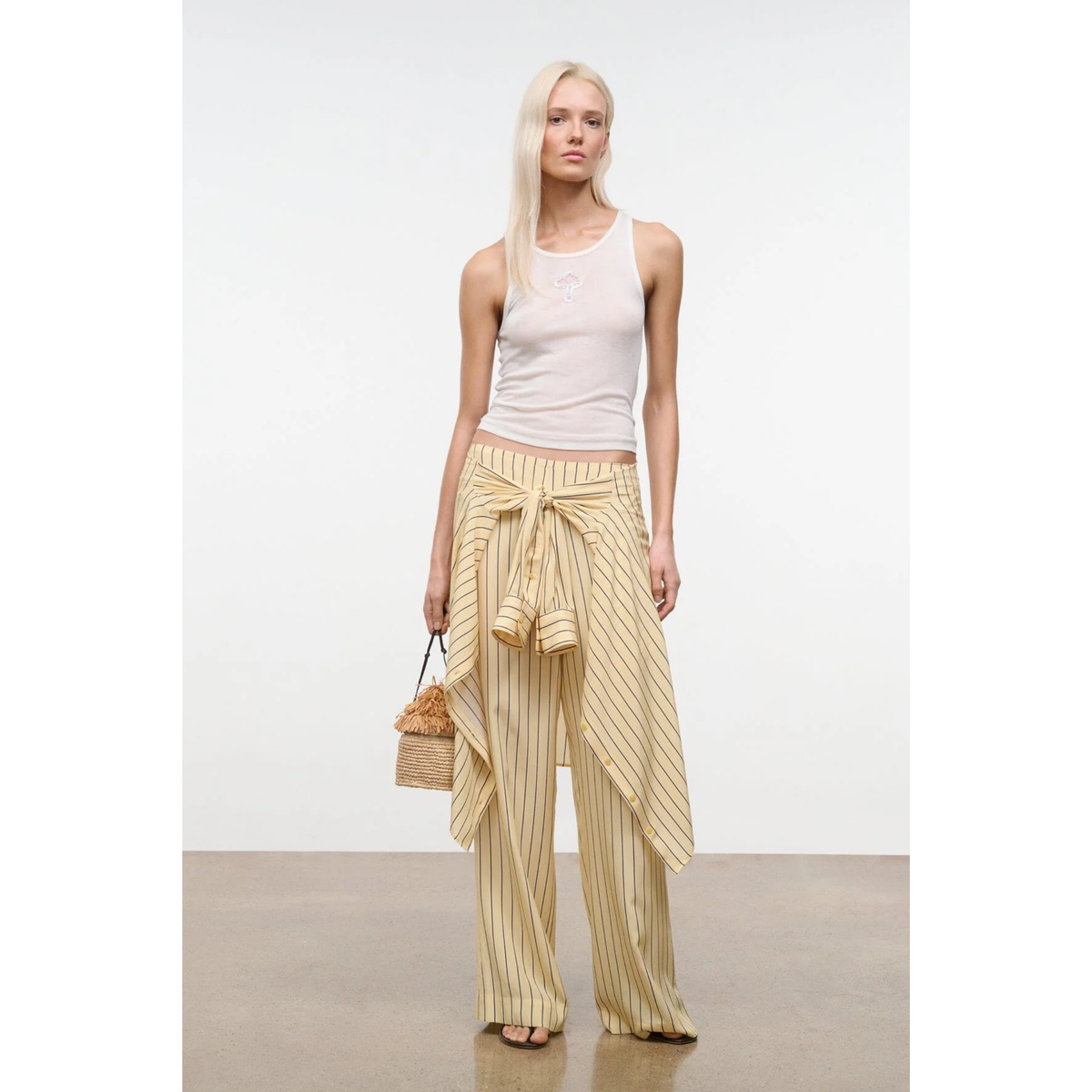 Woman wearing a light-colored sleeveless top and striped pants, holding a woven bag on a plain background.