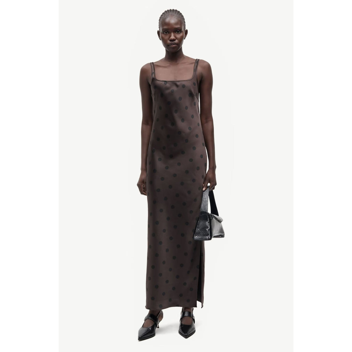 A model wearing a dark brown, floor-length slip dress with a small black polka dot pattern, holding a black handbag and wearing black pointed-toe shoes against a plain off-white background.
