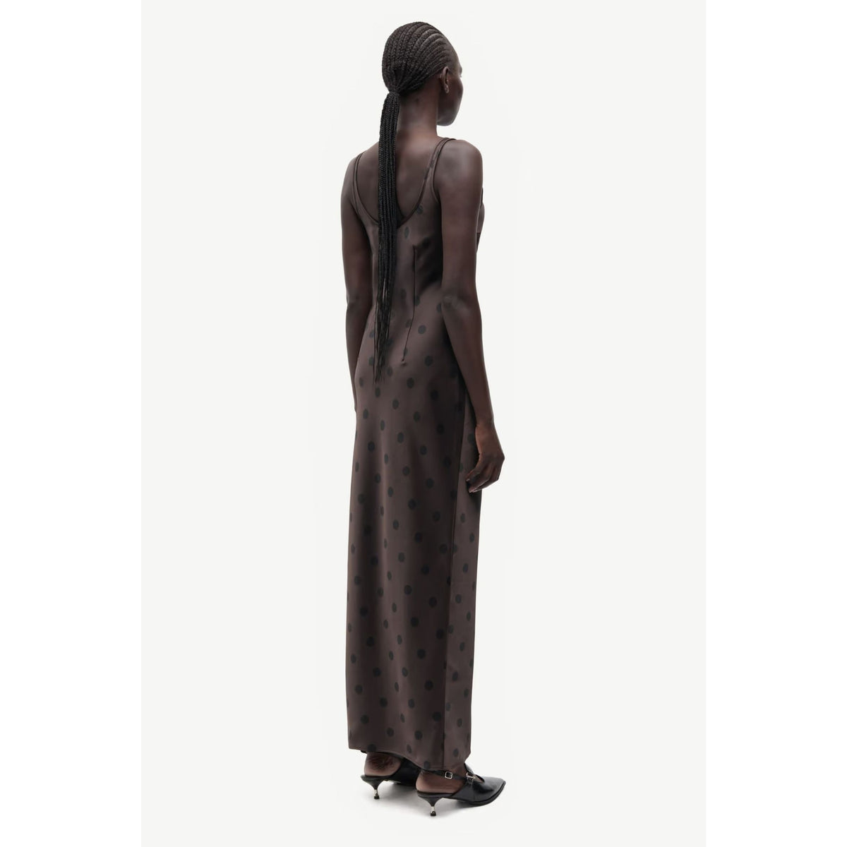 A model shown from the back wearing a dark brown sleeveless maxi dress with a subtle polka dot pattern and a low open back, paired with black pointed-toe heels, against a light gray studio background.