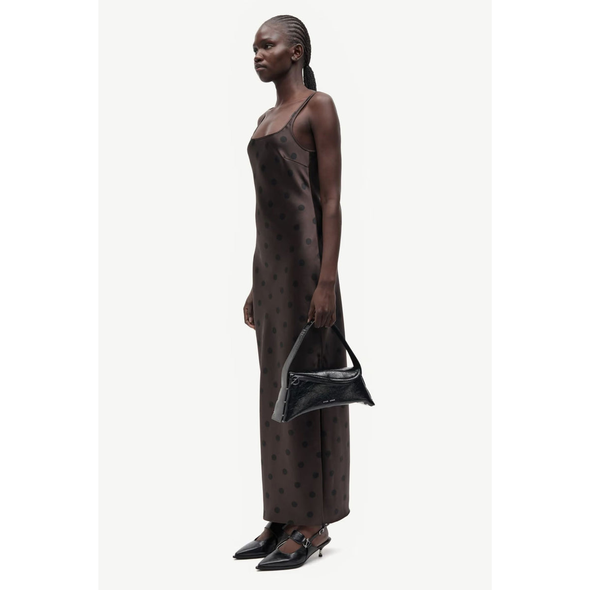 A model is shown in profile wearing a dark brown satin slip dress with a small polka dot pattern, holding a black handbag and wearing black pointed-toe flats against a light gray background.
