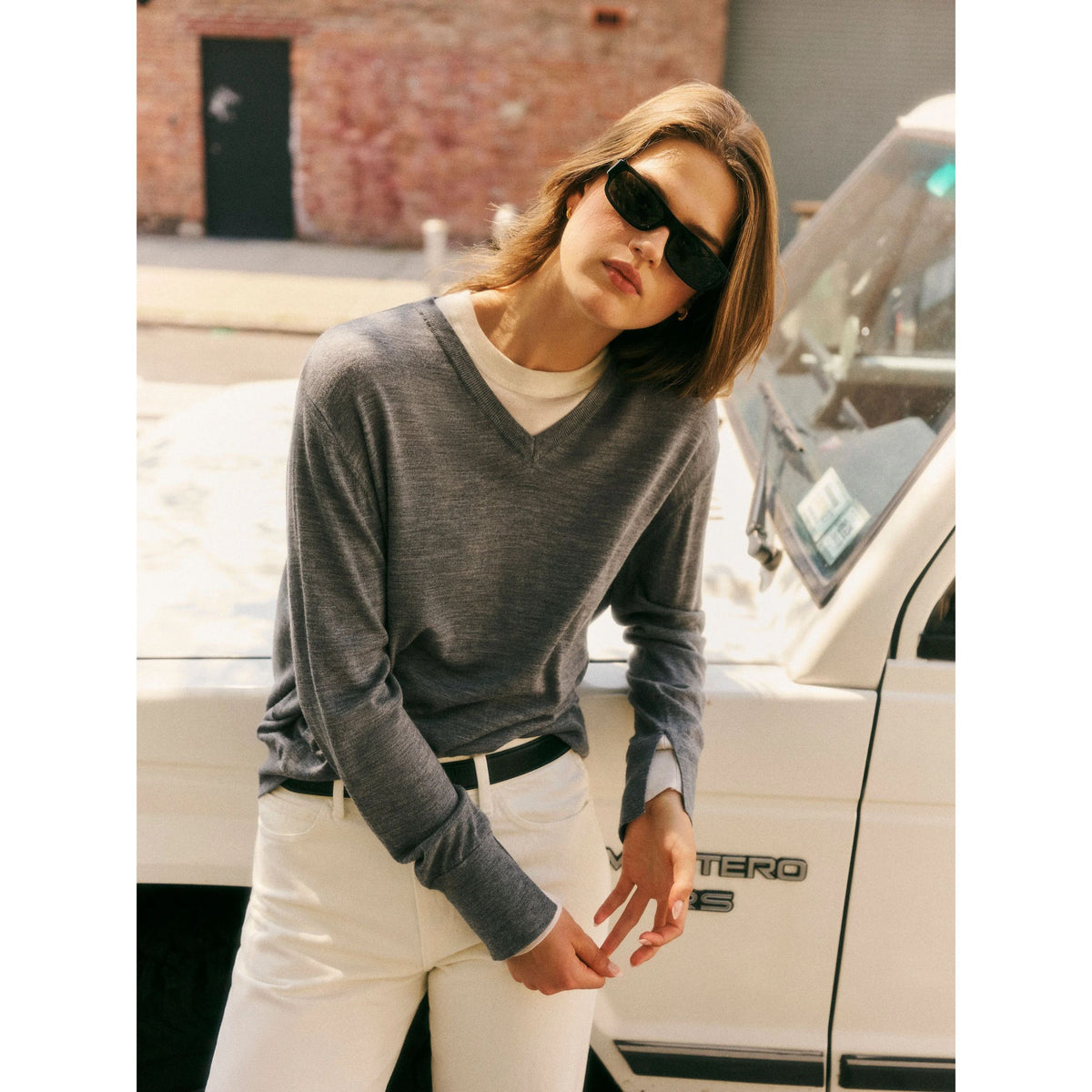 Medium shot of a model wearing sunglasses and the grey layered v-neck sweater with white pants, leaning against a white vehicle outdoors.