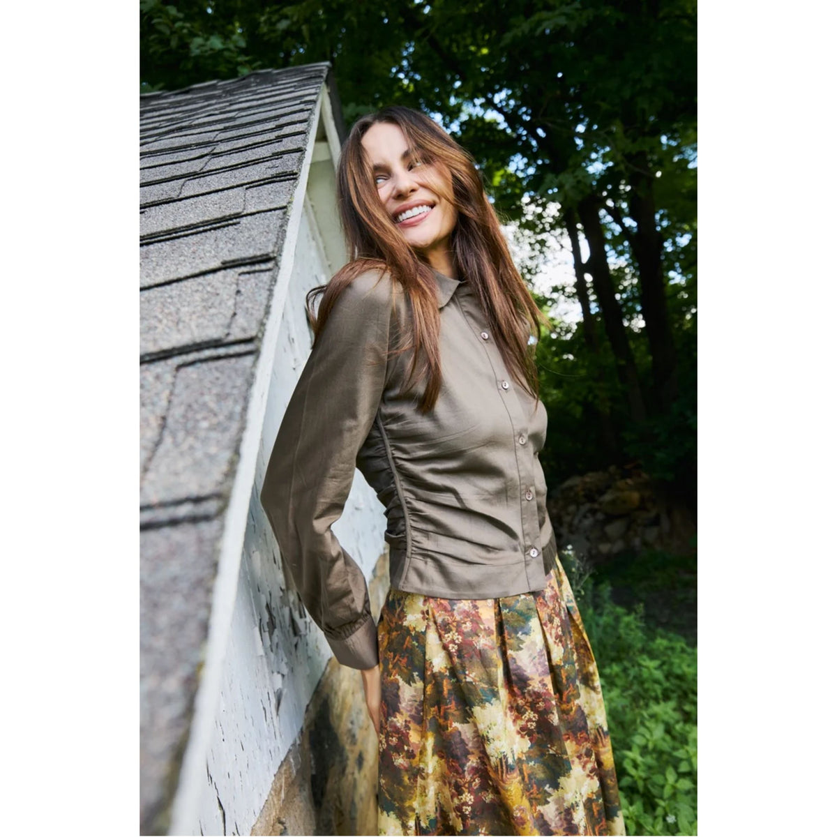 A woman wearing a muted olive-brown long-sleeve button-up shirt and a multicolor floral print skirt, leaning against a white wall next to a shingled roof with a forest background.