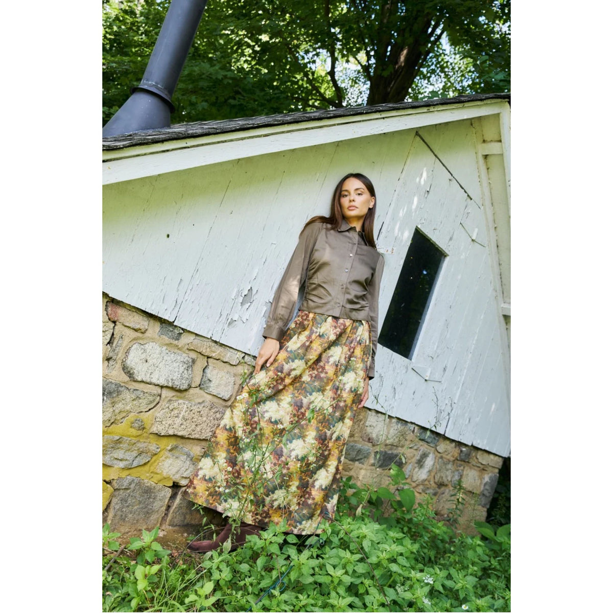 A woman stands outdoors in front of a weathered white wooden shed with a stone base, wearing a brown button-up shirt and a long floral print skirt.