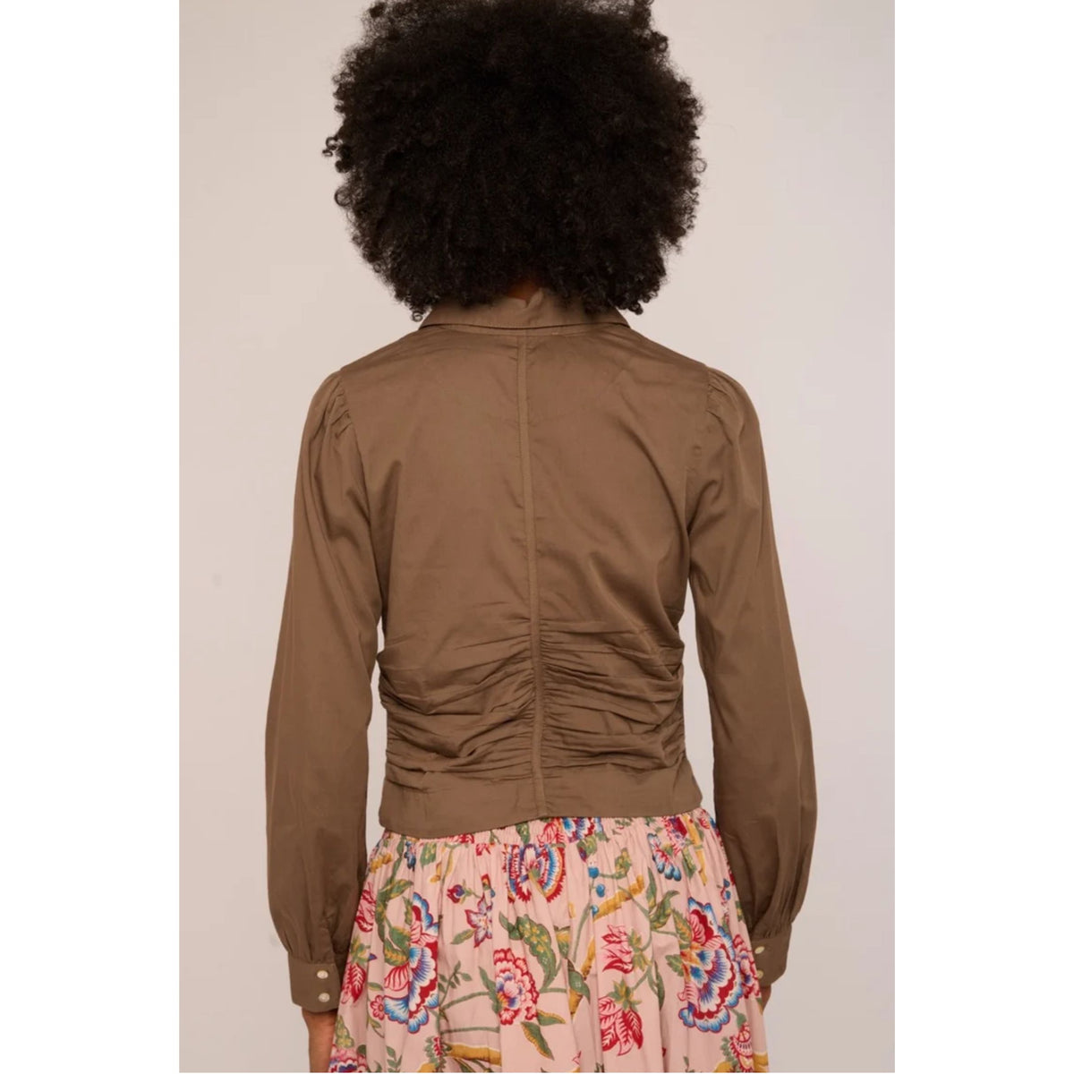 A person shown from the back wearing a cropped brown jacket with ruching down the center back and a floral skirt, against a plain light background.