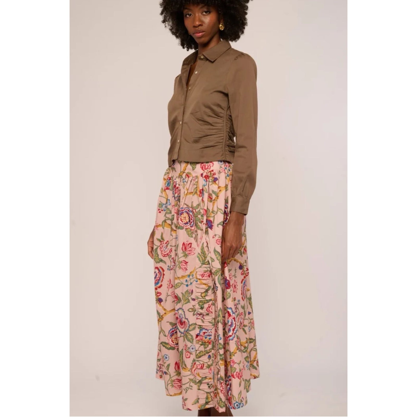 A woman with a large black afro wearing a cropped, long-sleeve brown button-up shirt and a pink floral skirt, standing against a plain light gray background.