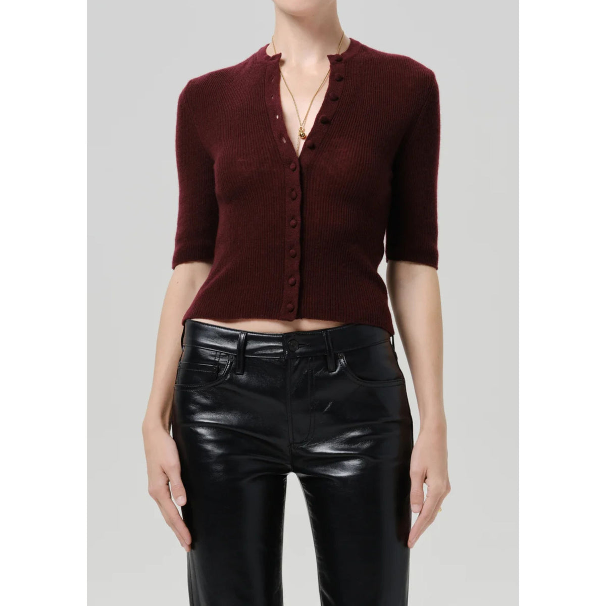 A model wearing a short-sleeve ribbed knit cardigan in deep burgundy with a buttoned split neckline, paired with black faux-leather pants, against a light gray studio background.
