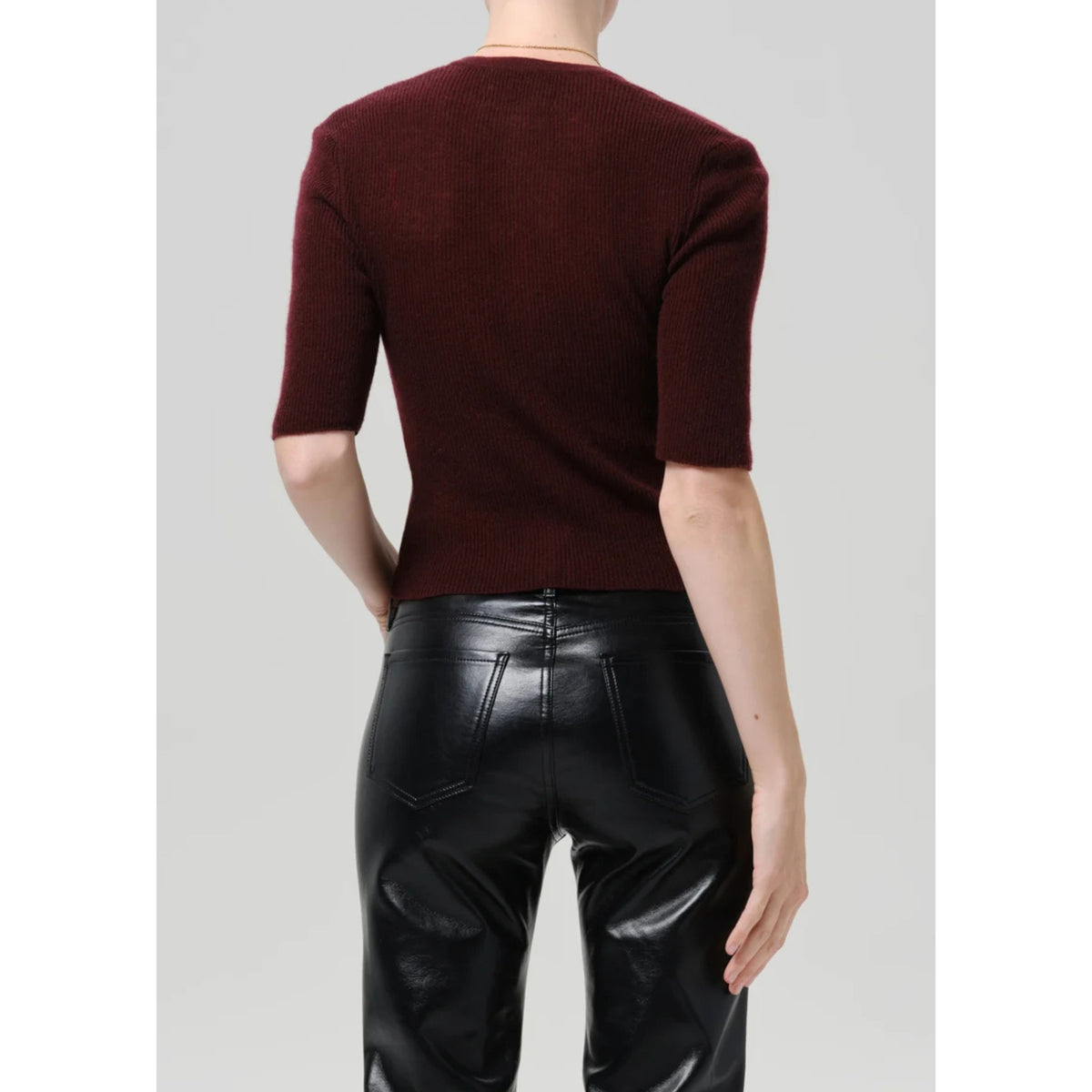 A model shown from the back wearing a short-sleeve burgundy knit top and shiny black faux-leather pants, set against a plain light gray studio background.
