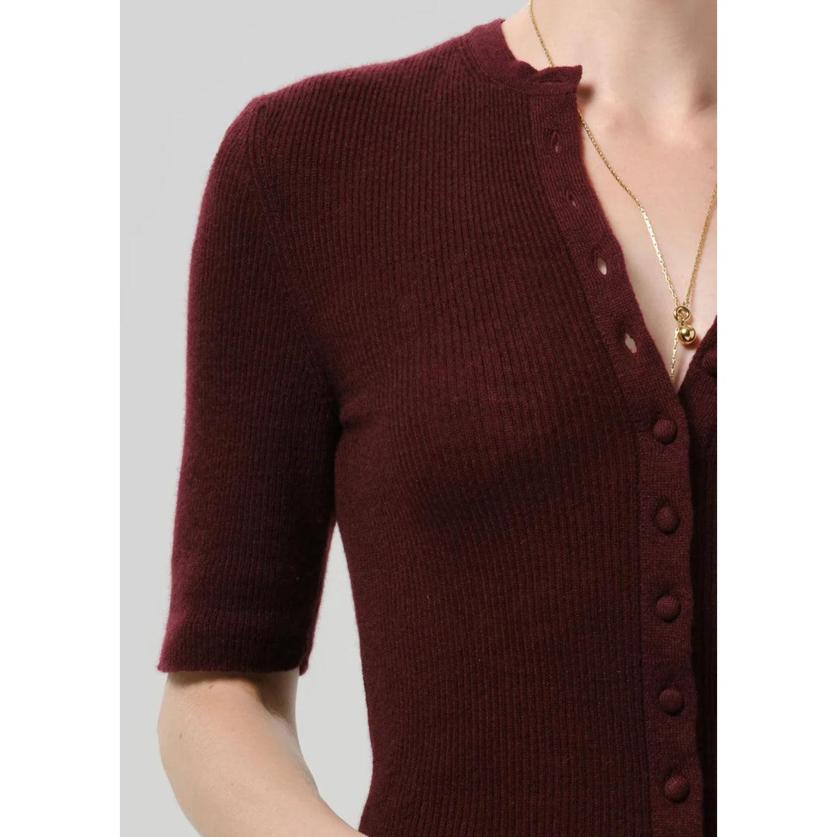 A close-up of a woman wearing a ribbed short-sleeve cardigan in deep burgundy with a buttoned V-neckline, shown against a light gray background.