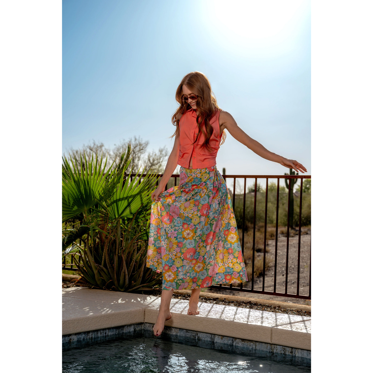 Woman in a colorful floral skirt and red top standing by a pool with desert landscape in the background