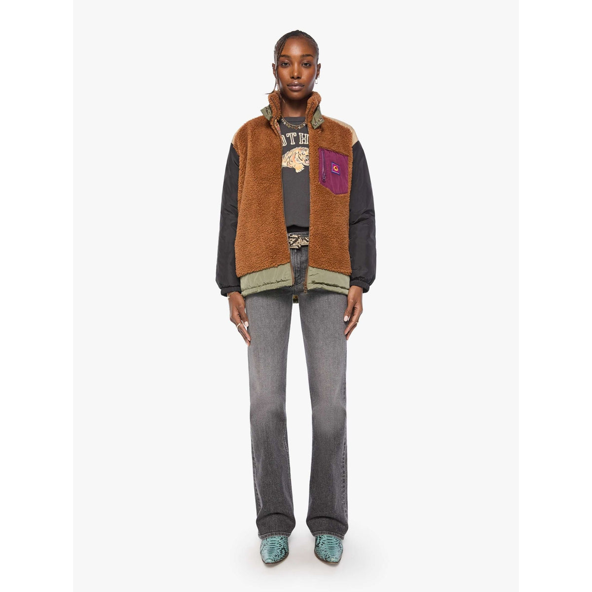 A model stands against a light gray studio background wearing a brown sherpa-lined patchwork jacket with dark sleeves, gray jeans, and teal shoes.