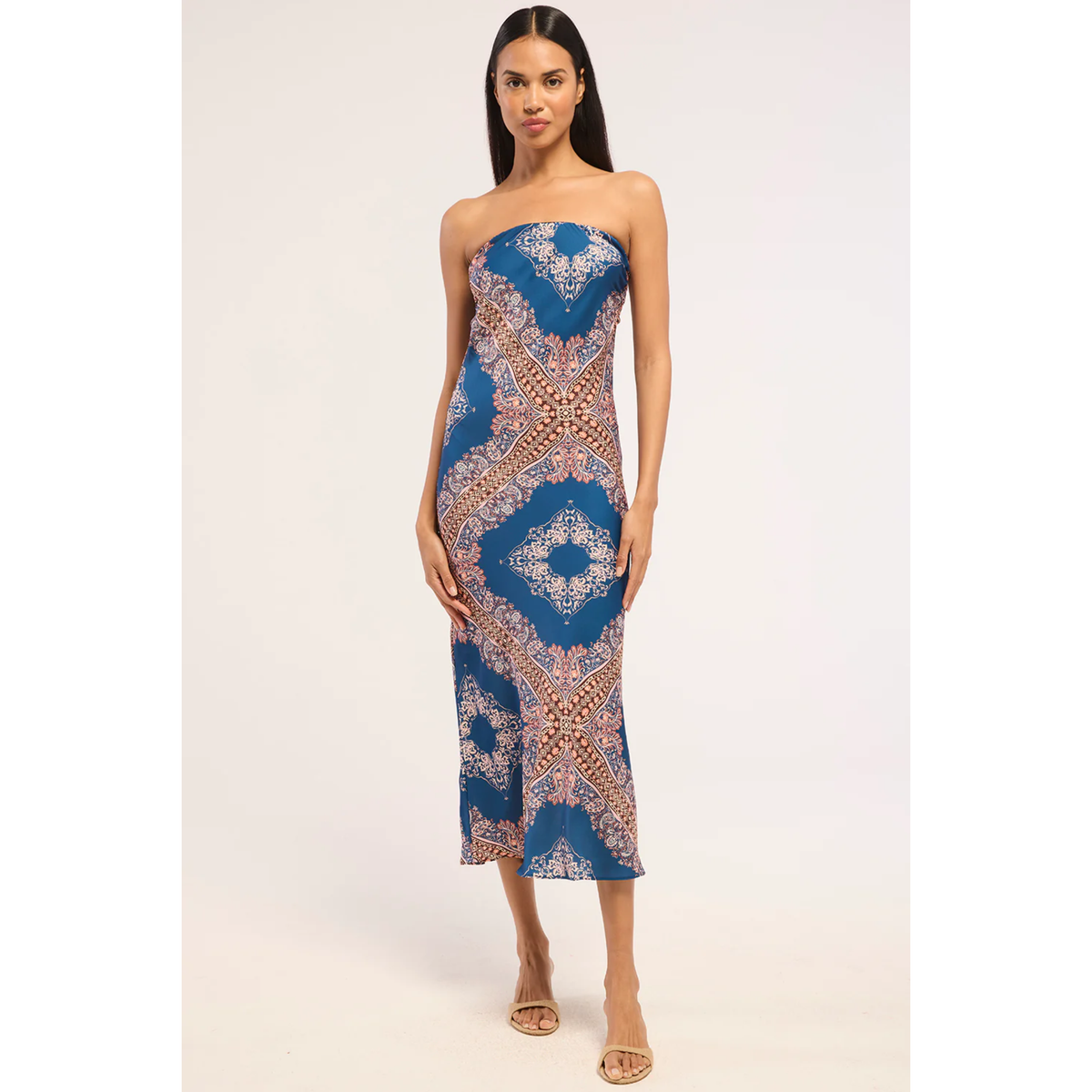 A woman modeling a strapless midi dress with a blue base and intricate white, beige, and orange geometric mandala patterns, set against a plain light background.