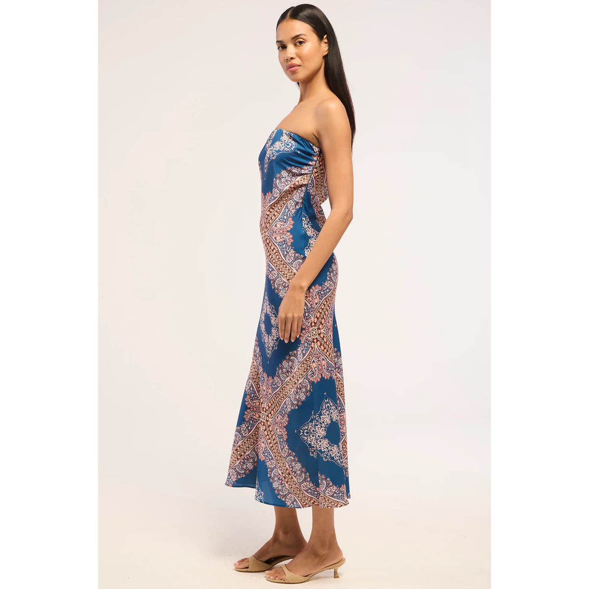 A woman modeling a strapless midi dress with a blue base and a beige paisley pattern, standing against a plain light background.