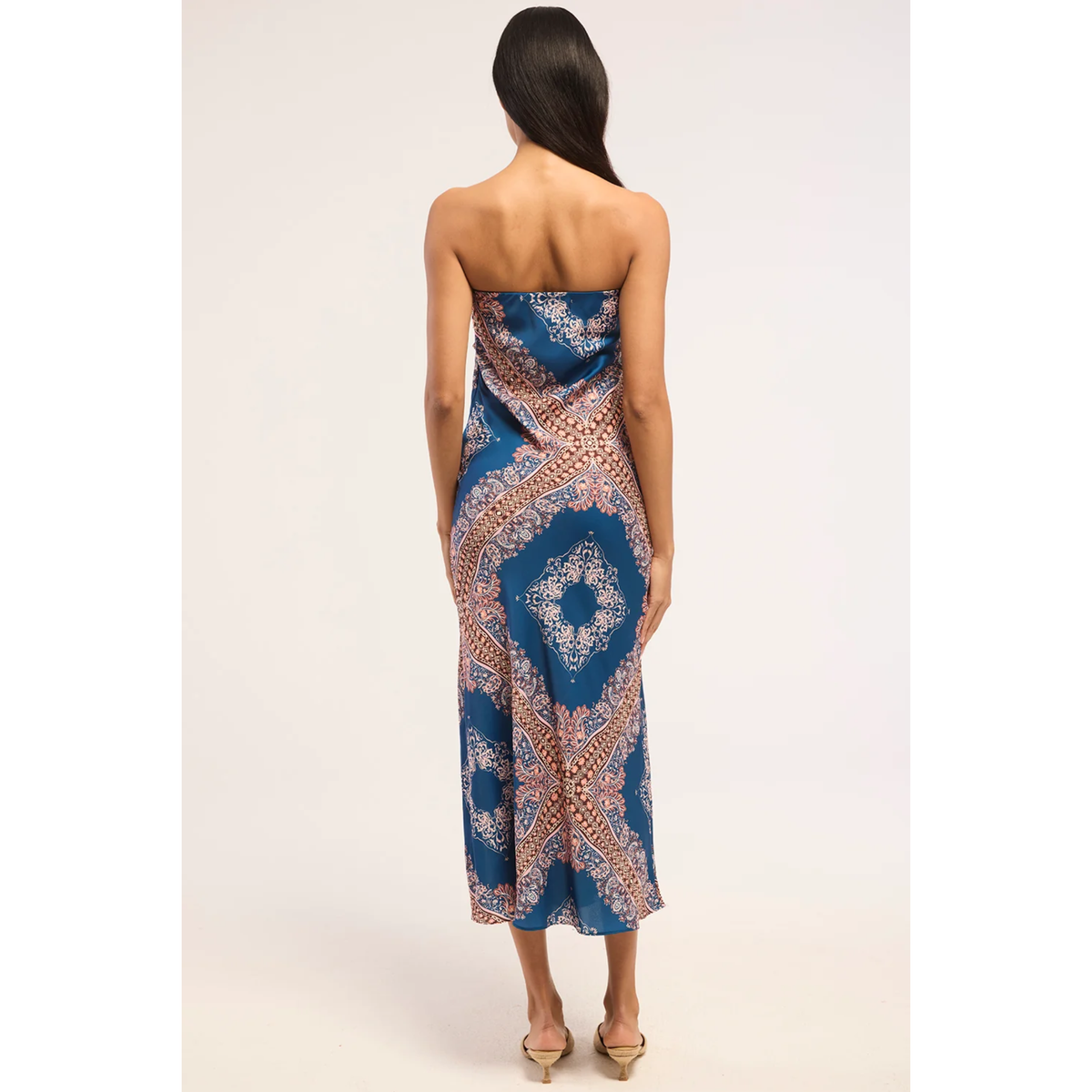 A woman viewed from the back wearing a strapless midi dress with a blue, beige, and brown geometric pattern, set against a plain light background.