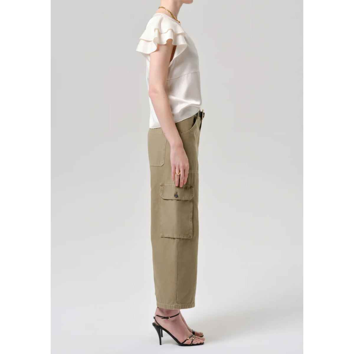 A model shown from the side wearing a cream ruffled short-sleeve top and olive green cargo pants with black high-heeled sandals, set against a plain light gray studio background.