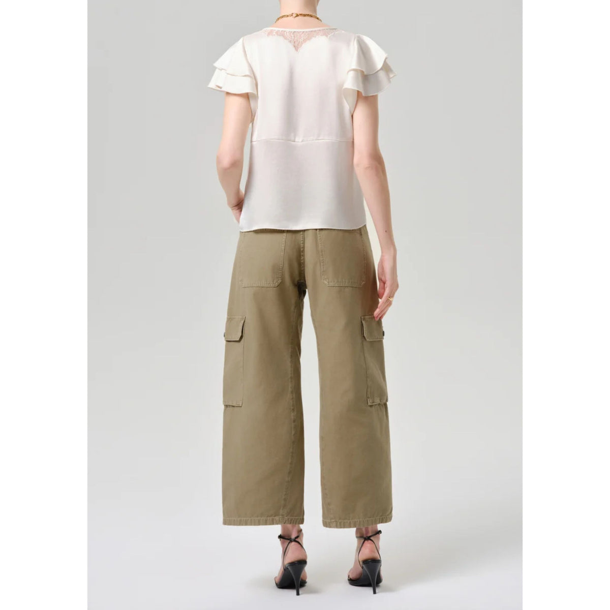 A model shown from the back wearing a cream-colored ruffled short-sleeve blouse and olive green wide-leg cargo pants, paired with black high-heeled sandals, against a light gray studio background.