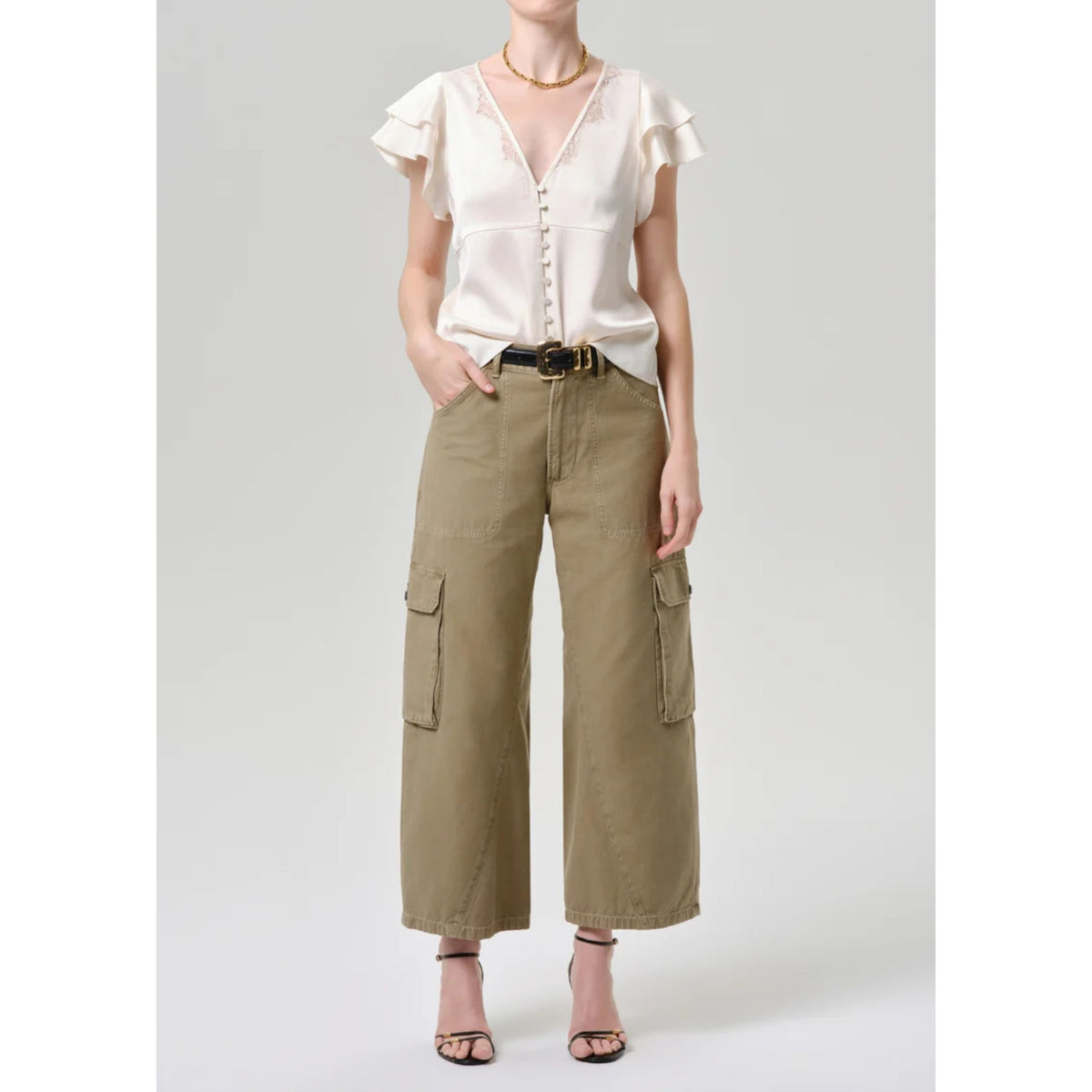 A model wearing a cream ruffled short-sleeve blouse and olive green wide-leg cargo pants, styled with a black belt and black strappy heels against a light gray studio background.