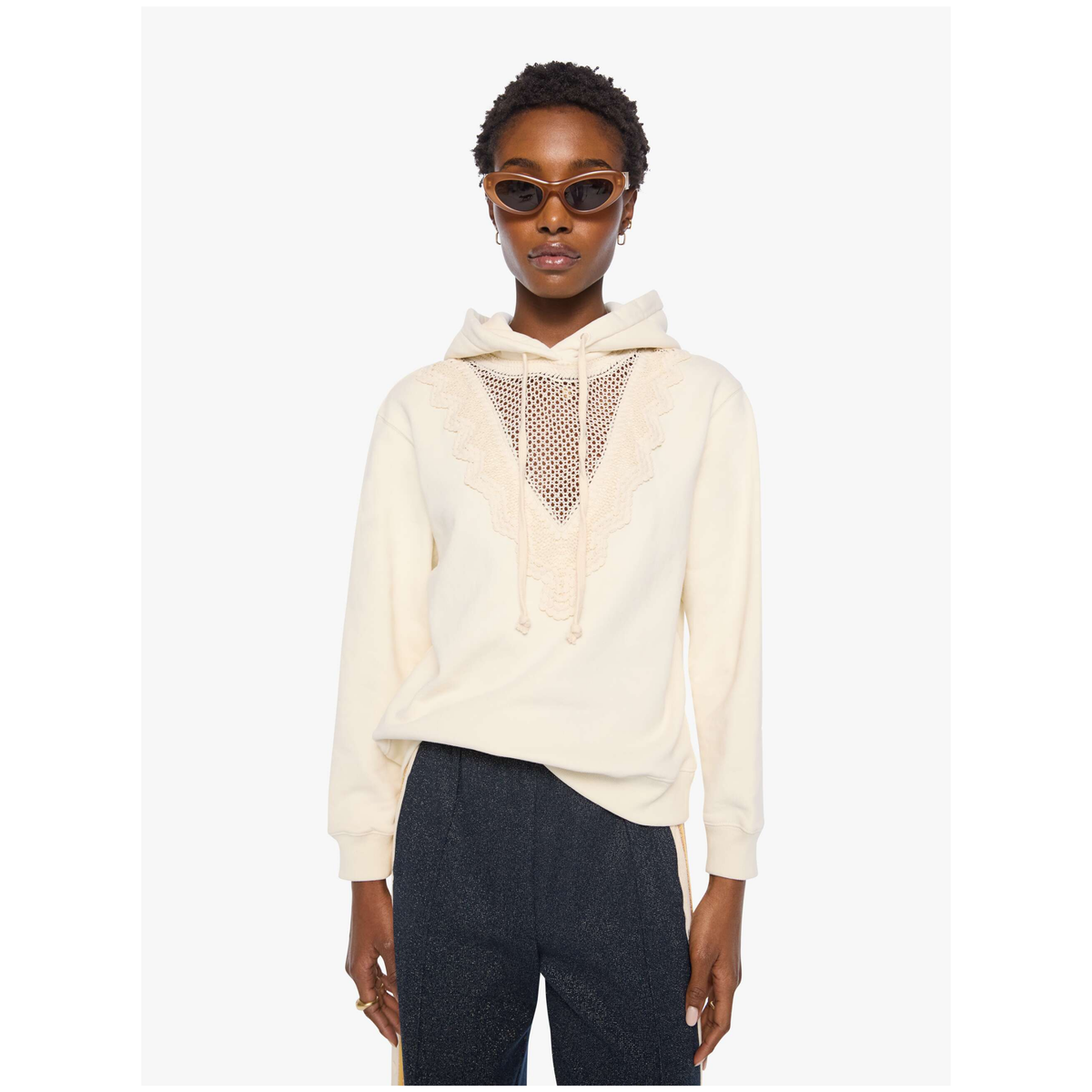 A model wearing a cream-colored hooded sweatshirt with a sheer mesh chest panel and dark charcoal pants, set against a plain light gray background.