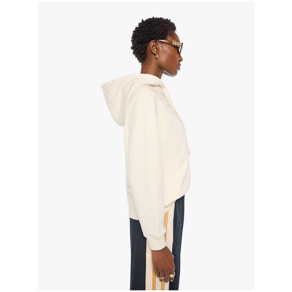 A model shown from the side profile wearing a cream-colored cropped hoodie and dark pants with gold stripes, set against a plain light gray background.