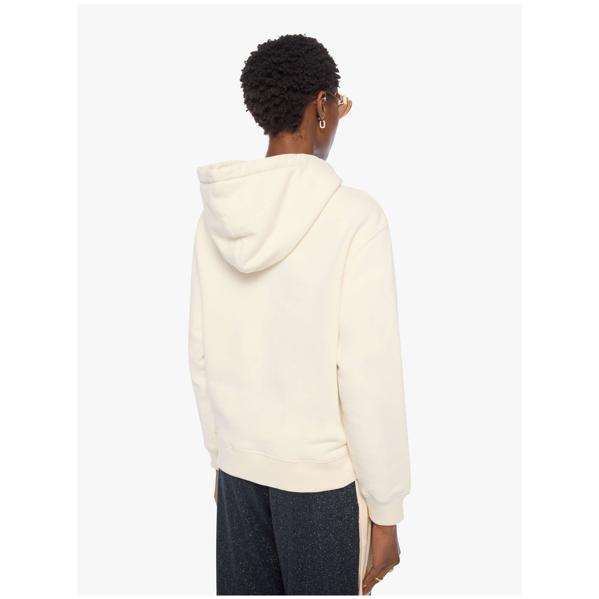 A person shown from the back wearing a cream-colored hooded sweatshirt and dark pants against a light gray studio background.