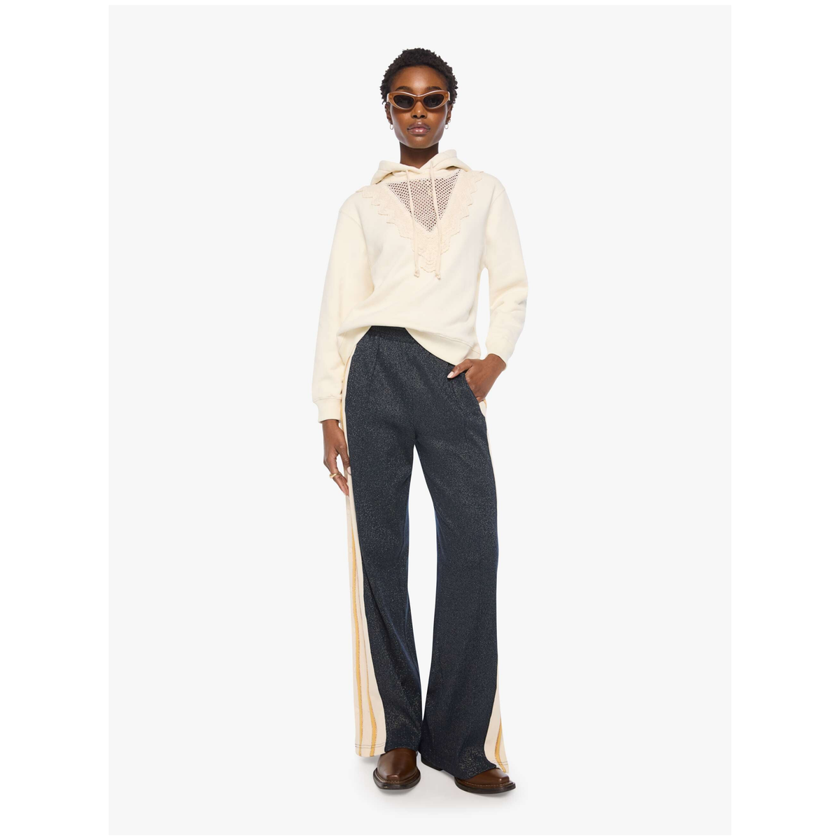 A model wearing a cream-colored lace-trimmed long-sleeve top and dark blue wide-leg trousers with beige side stripes, standing against a plain white studio background.