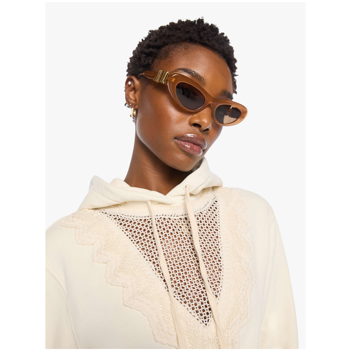 A model wearing a cream-colored hoodie with a textured crochet chest panel and lace trim, styled with cat-eye sunglasses against a plain white background.