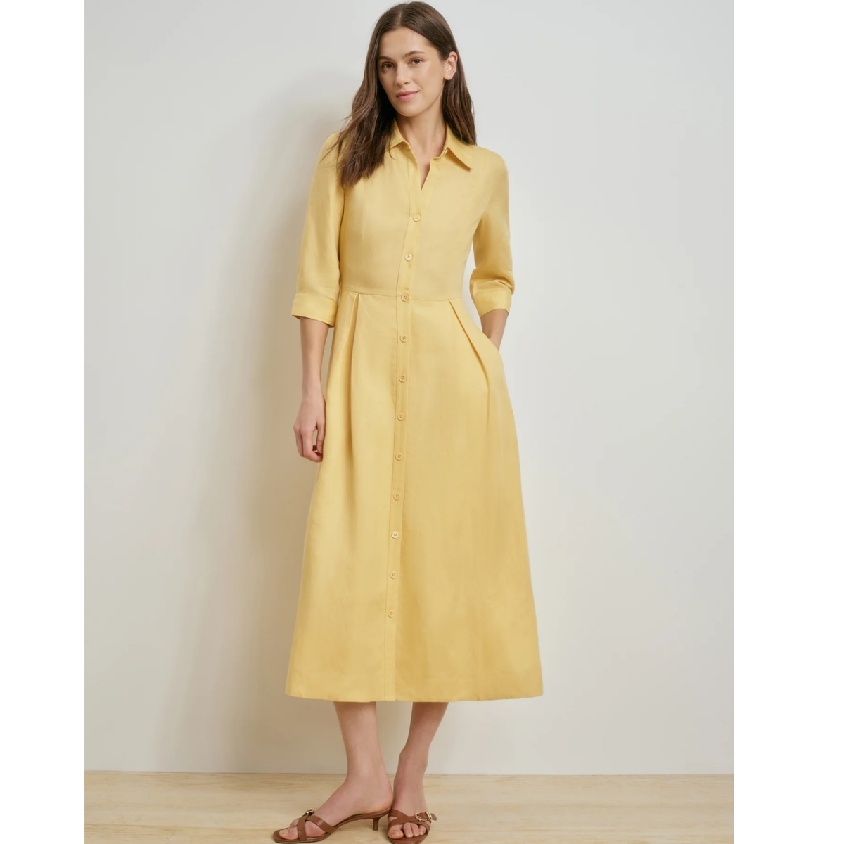 A woman modeling a light yellow linen shirt dress with three-quarter sleeves, a button-down front, and a gathered waist, paired with brown flat sandals against a neutral studio background.