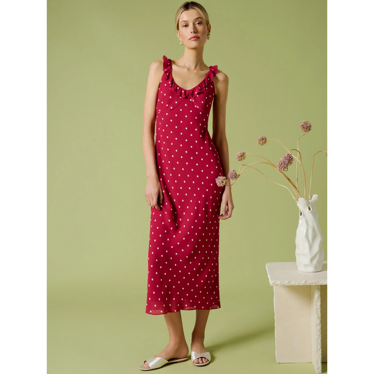 Woman wearing a red polka dot dress standing against a green background with a decorative vase.