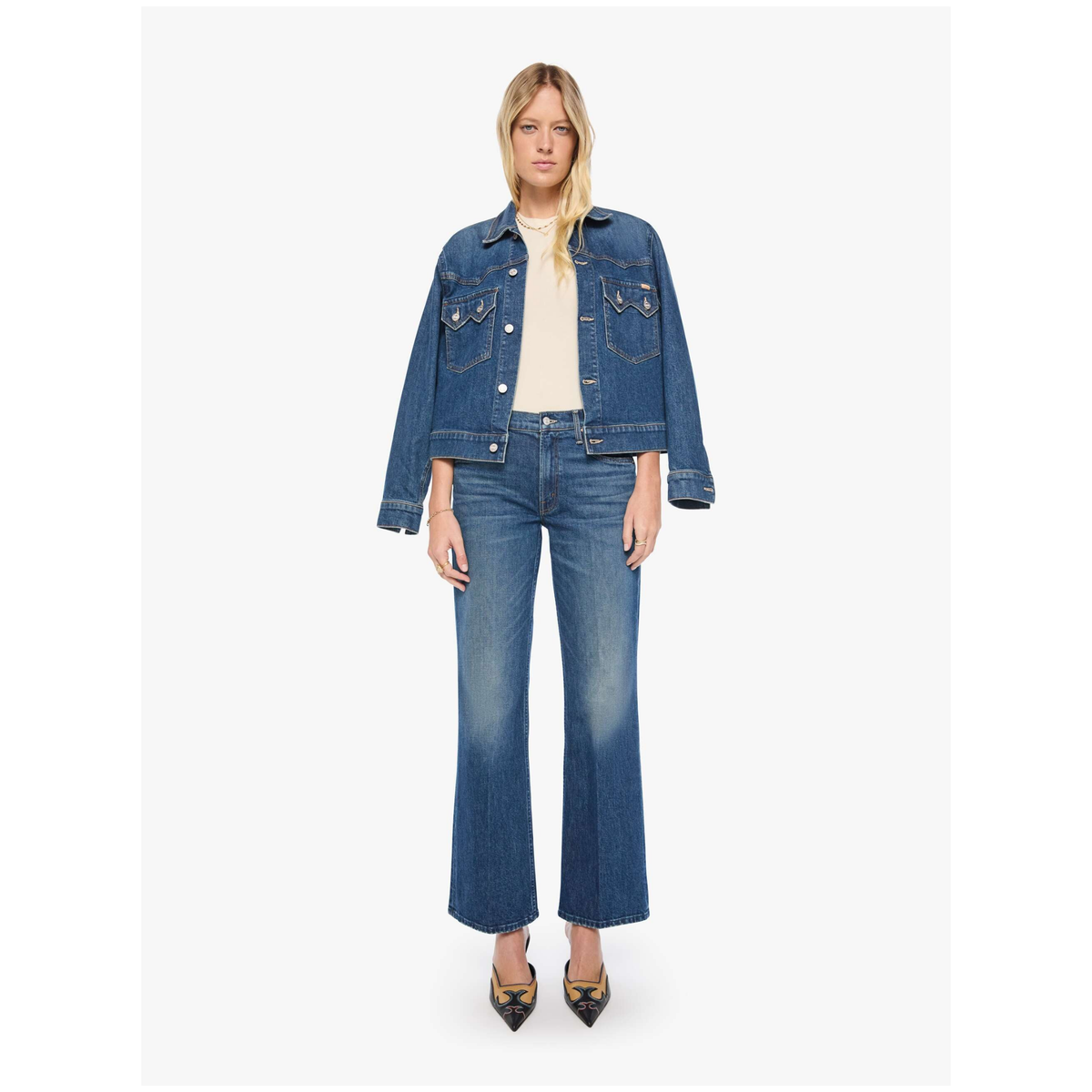 A model stands against a light gray studio background wearing a blue denim jacket and matching blue denim flare jeans over a cream top, with black pointed heels.