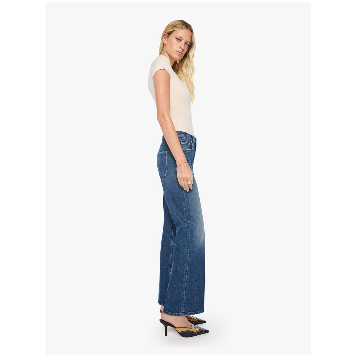 A woman modeling a pair of wide-leg blue denim jeans with a cream short-sleeve top and black pointed-toe heels, shown in profile against a light gray studio background.