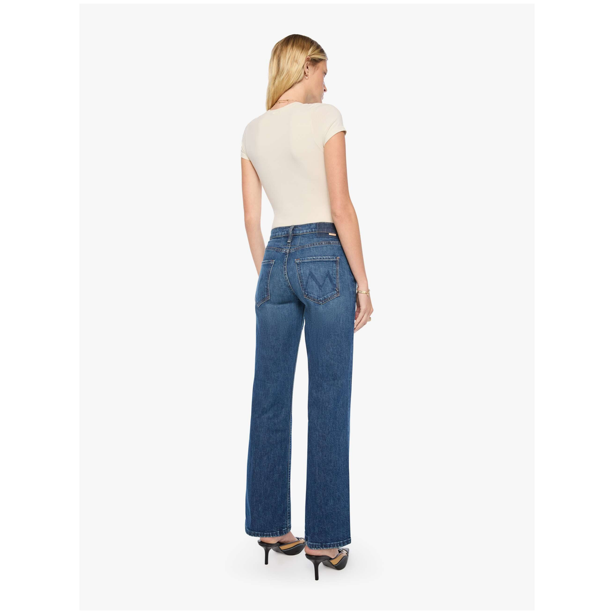 A woman shown from the back wearing blue wide-leg jeans, a cream short-sleeve top, and black high heels against a light gray studio background.