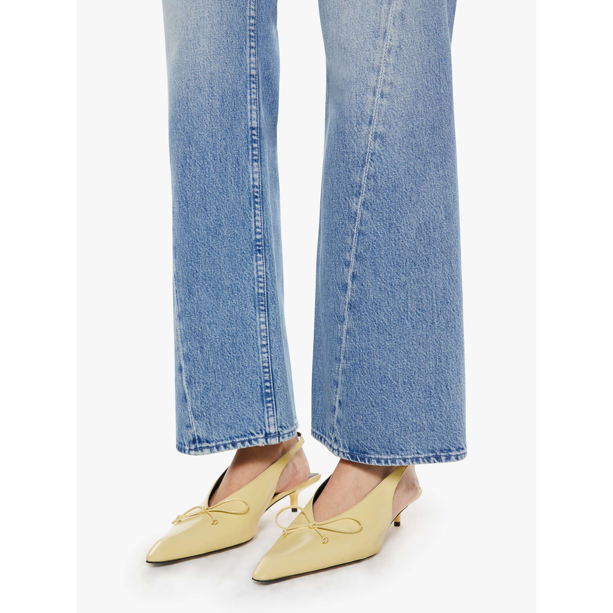 A person wearing light blue flared jeans and pale yellow pointed-toe mule heels with bow details, shown from the calves down on a clean white studio background.