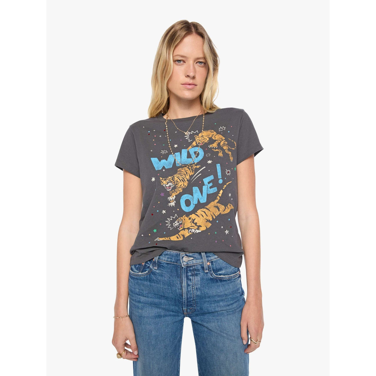 A woman modeling a dark gray graphic t-shirt with a tiger and the words 'WILD ONE!' in blue and orange, paired with blue jeans, against a light gray background.