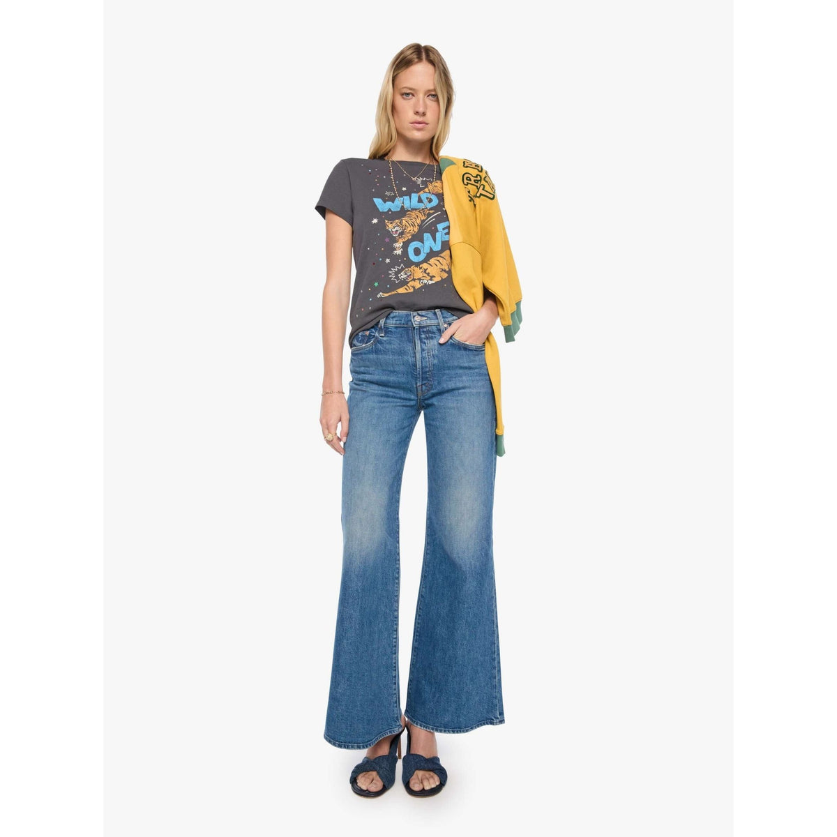 A woman modeling a dark gray graphic t-shirt with blue and yellow text, paired with high-waisted blue flare jeans and a yellow cardigan draped over her shoulder, set against a plain white studio background.