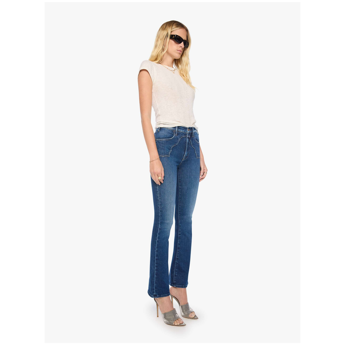 A woman modeling a pair of medium-wash blue flare jeans with a white sleeveless top and gray high-heeled sandals against a light gray studio background.