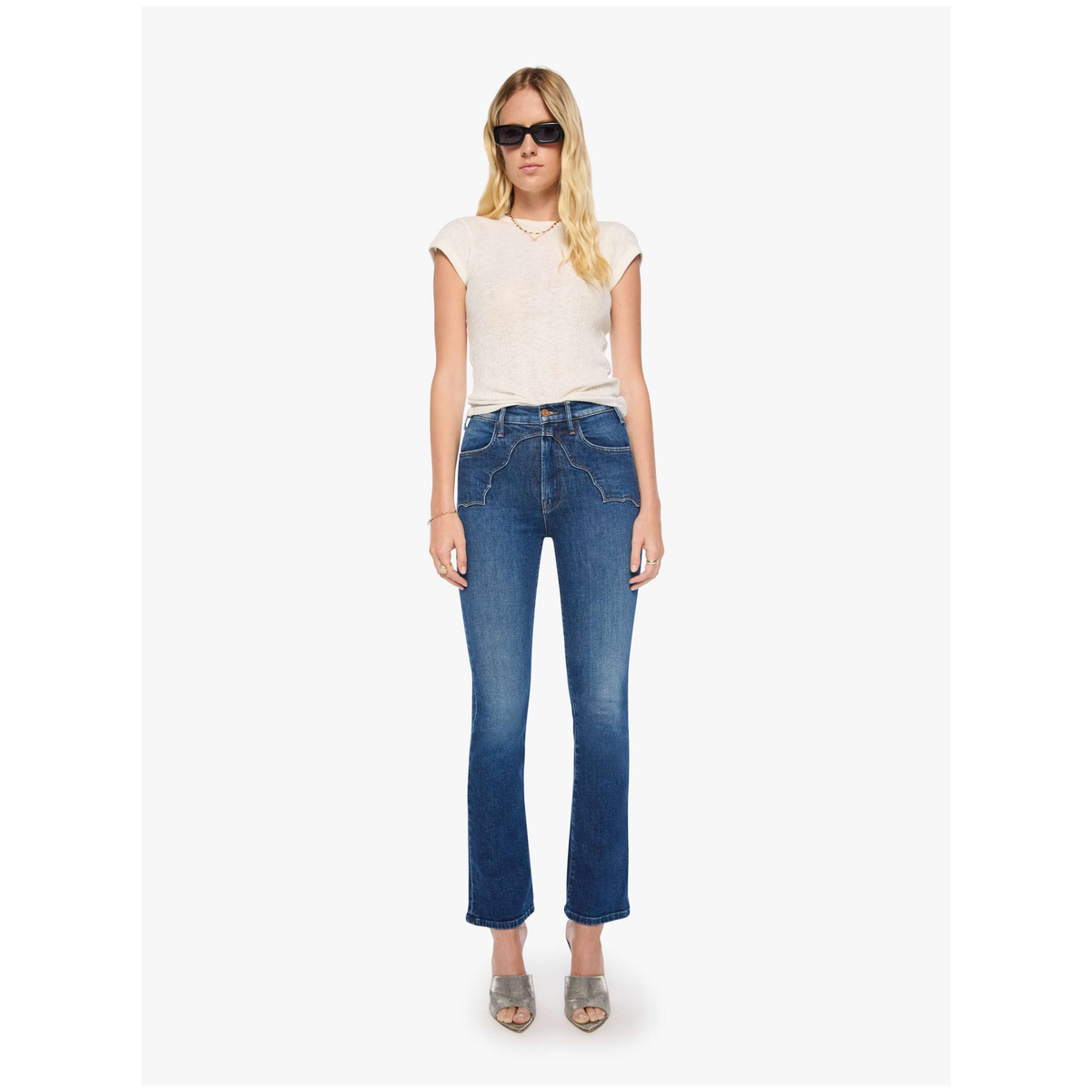 A woman modeling a pair of blue cropped flare jeans with a cream short-sleeve top and sunglasses against a plain white background.