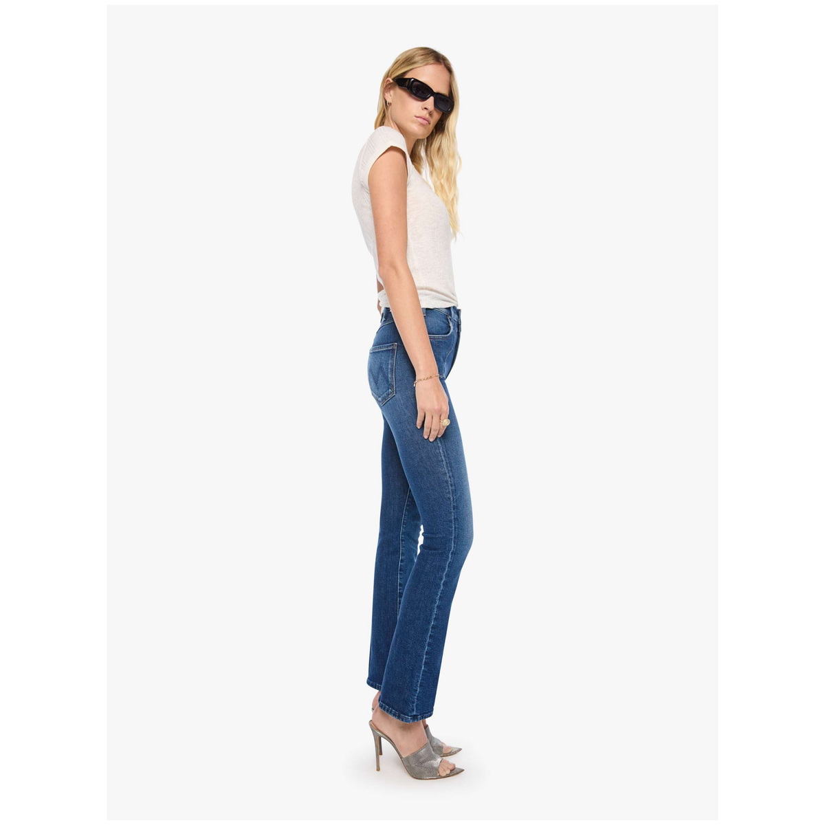 A woman modeling a pair of blue straight-leg jeans, paired with a white sleeveless top and grey high-heeled sandals, shown in profile against a plain white studio background.