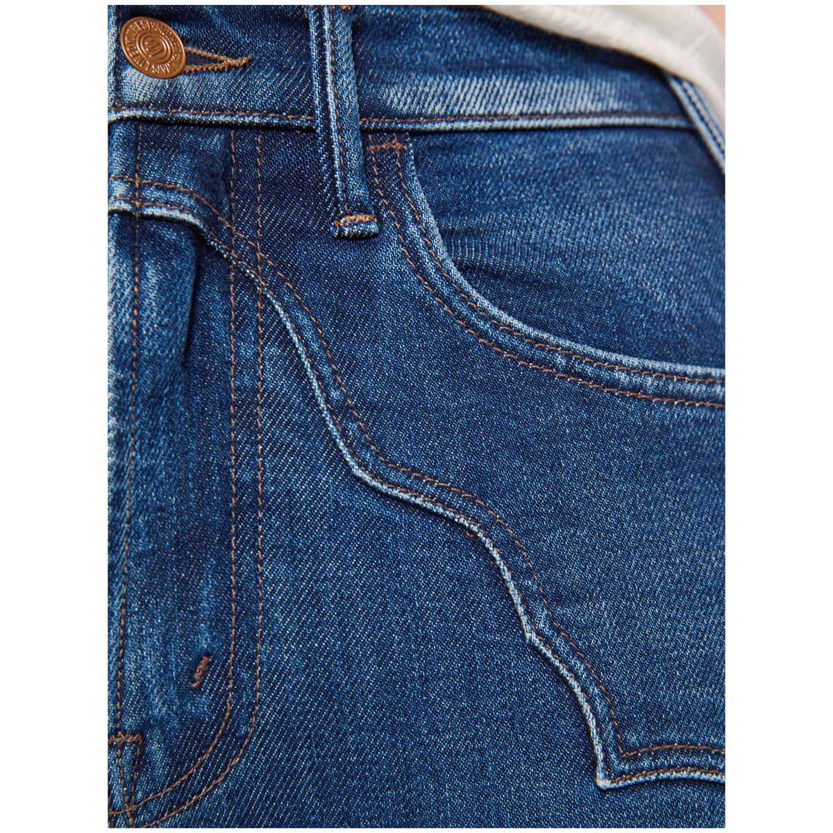 Close-up of the back pocket area of blue denim jeans, showing the blue denim fabric, brown contrast stitching, a brown metal button, and a small portion of a light-colored garment at the waistband.