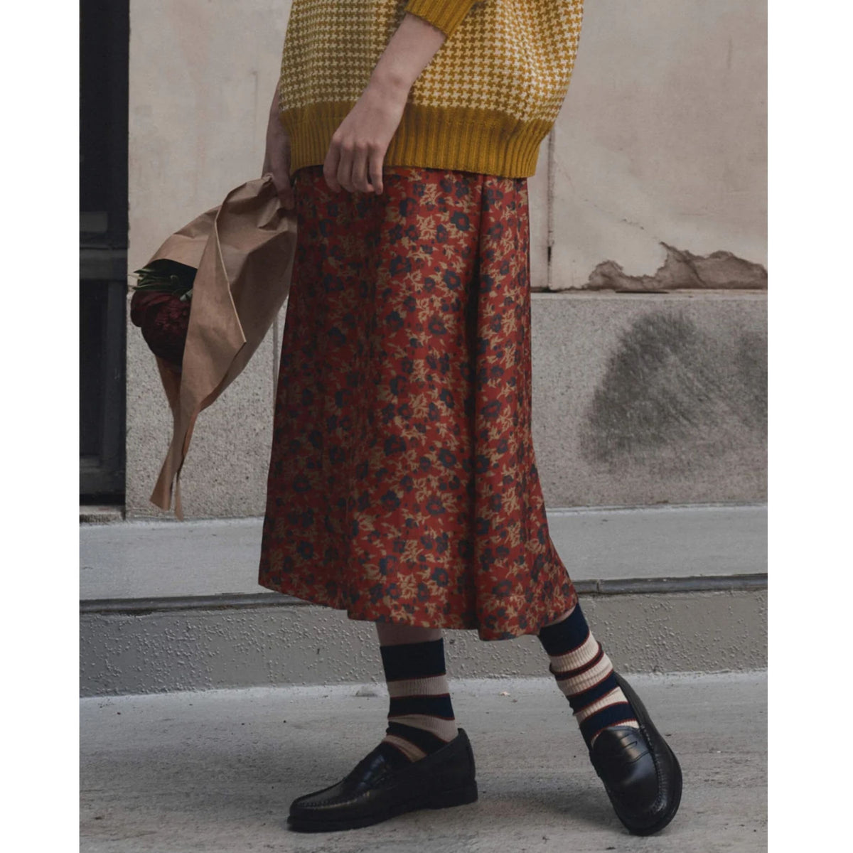 A person wearing a rust-red midi skirt with a dense floral print in orange, yellow, and blue, styled with a yellow knit sweater, striped socks, and black loafers, standing on a city sidewalk.