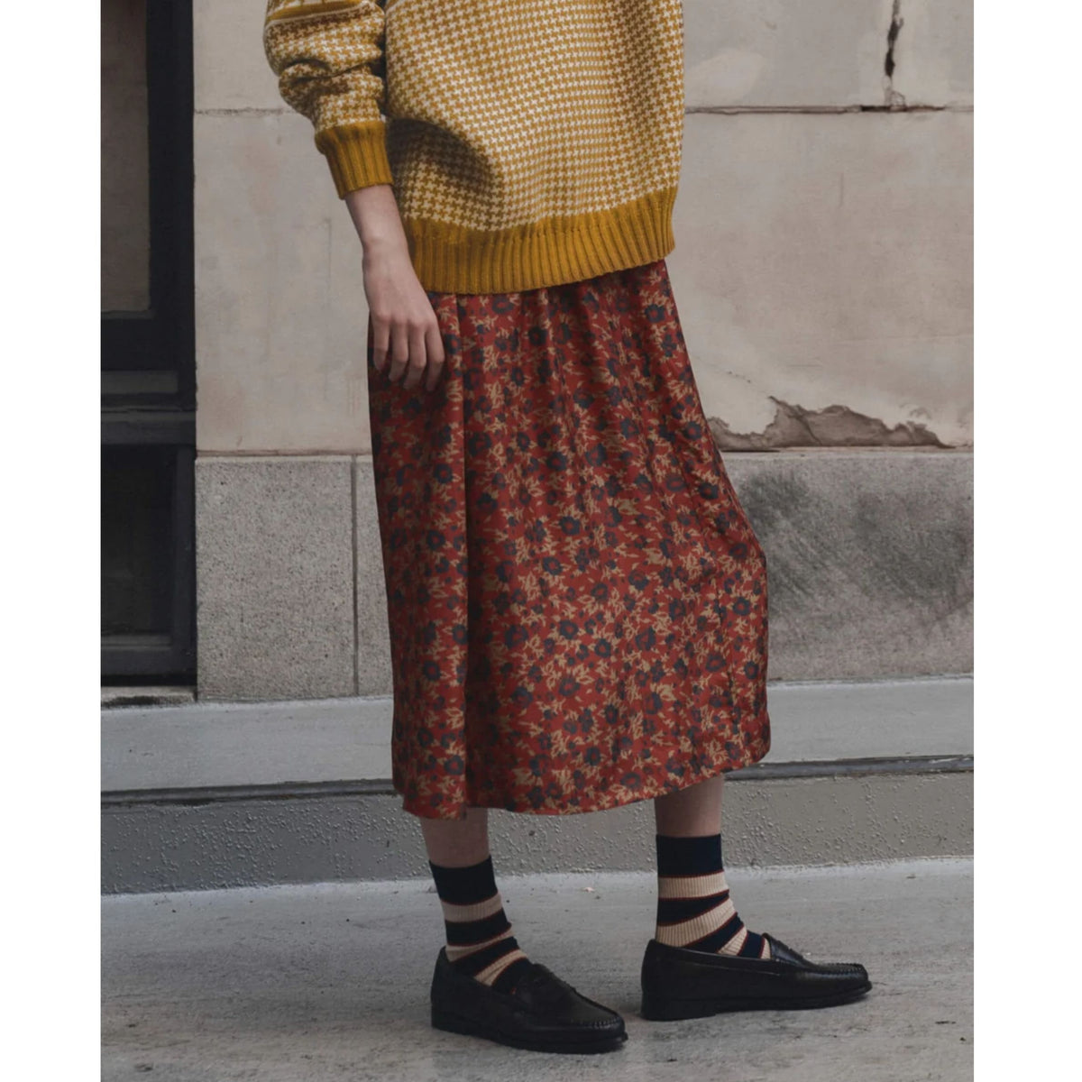 A person wearing a rust-red midi skirt with a dense small floral print, paired with a mustard yellow and white checkered sweater, striped socks, and black loafers, standing outdoors against a stone wall.
