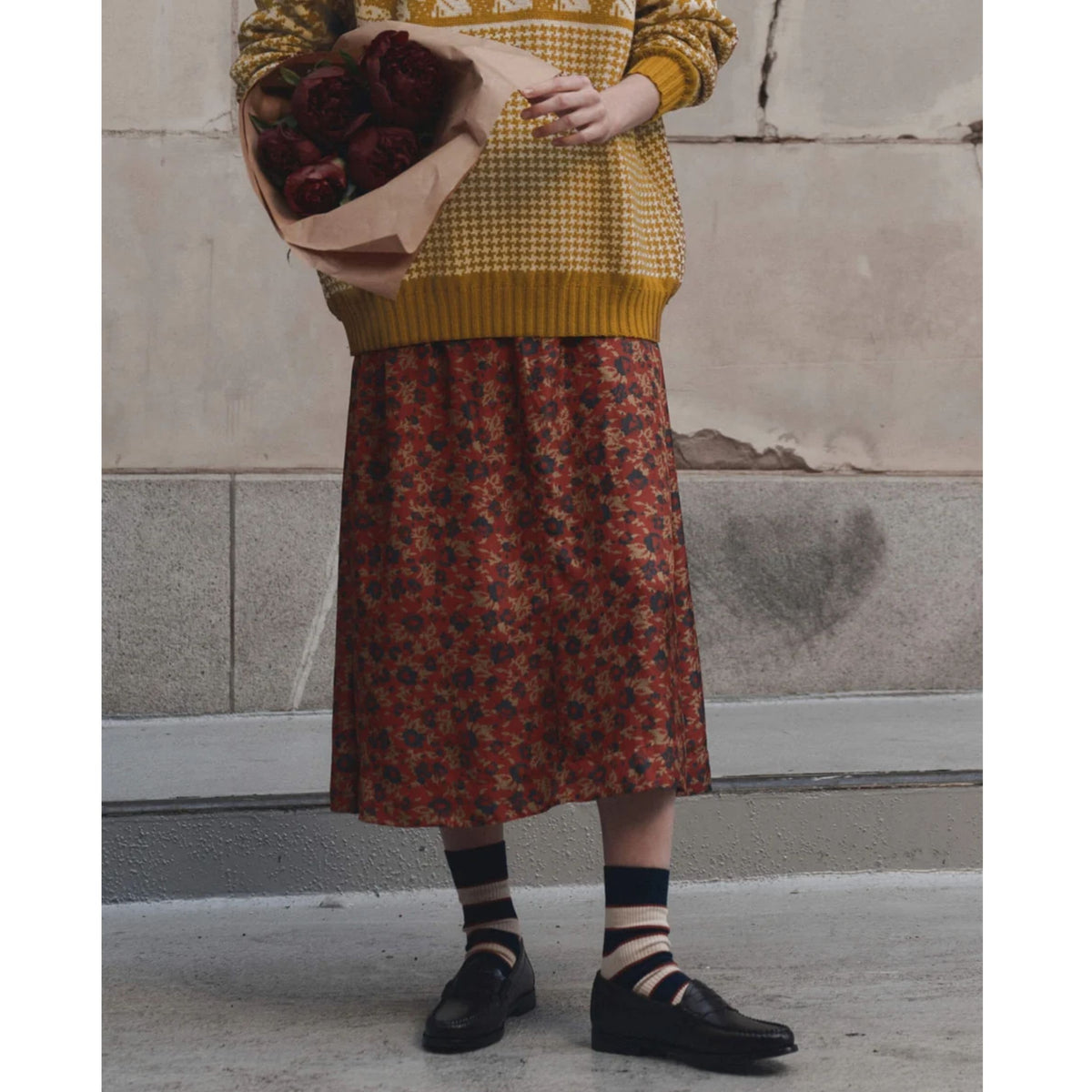 A person wearing a mustard yellow knit sweater and a rust-red midi skirt with a dark floral pattern, holding a paper bag of red roses, standing against a textured concrete wall.