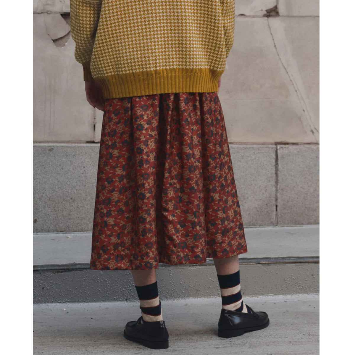 A person shown from the back wearing a mustard yellow houndstooth sweater and a rust-red midi skirt with a dense small floral print in blue and yellow. They are wearing black and beige striped socks and black Mary Jane shoes, standing against a concrete wall.